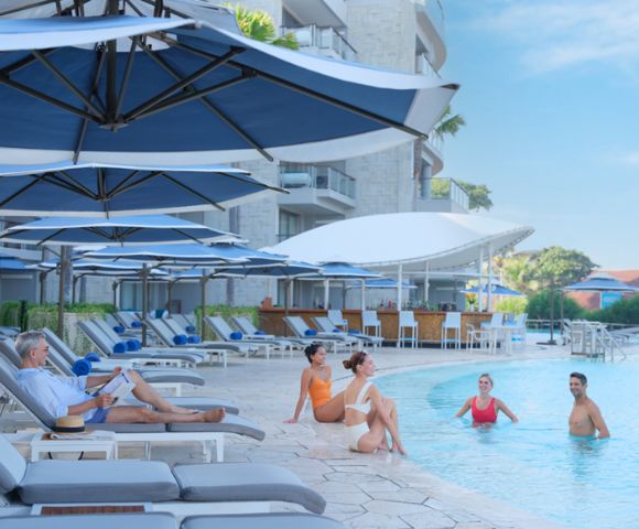 People enjoy a sunny day by a resort pool with loungers and large umbrellas. Some are seated poolside, others swim, surrounded by modern buildings.
