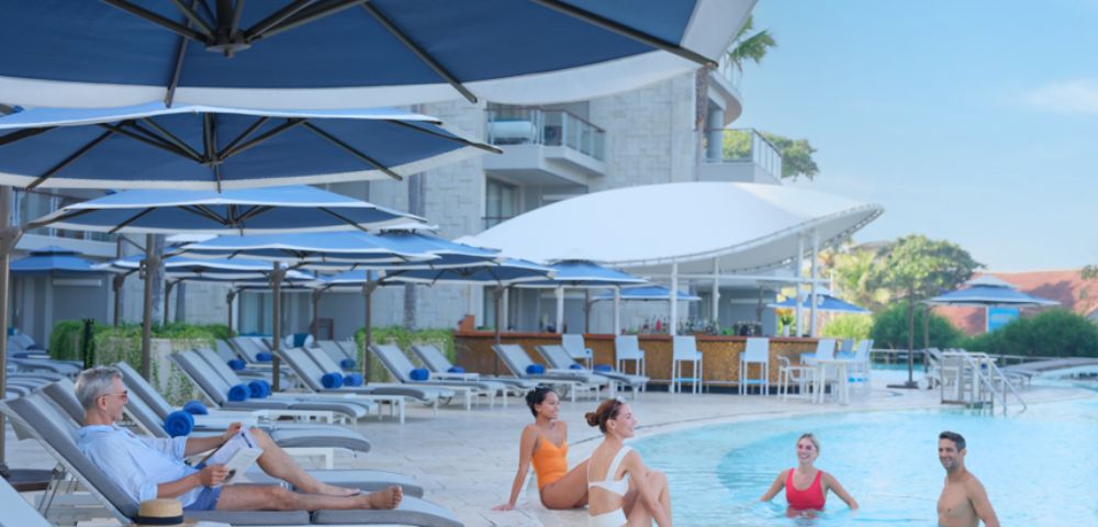 People enjoy a sunny day by a resort pool with loungers and large umbrellas. Some are seated poolside, others swim, surrounded by modern buildings.