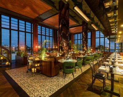 Luxurious restaurant interior with elegant green chairs, wooden tables, and warm lighting. Floor-to-ceiling windows provide city night views, enhancing the cozy ambiance.