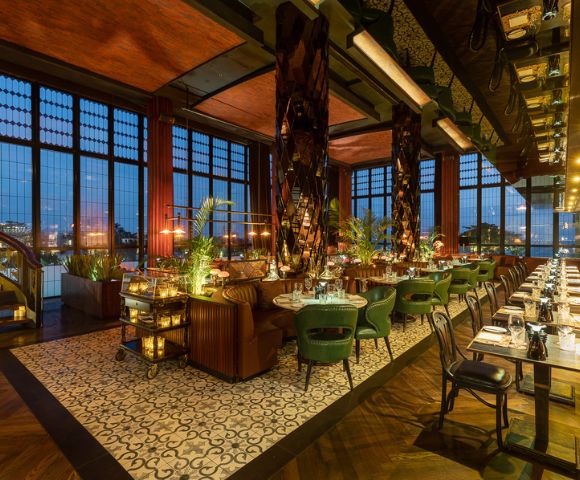 Luxurious restaurant interior with elegant green chairs, wooden tables, and warm lighting. Floor-to-ceiling windows provide city night views, enhancing the cozy ambiance.