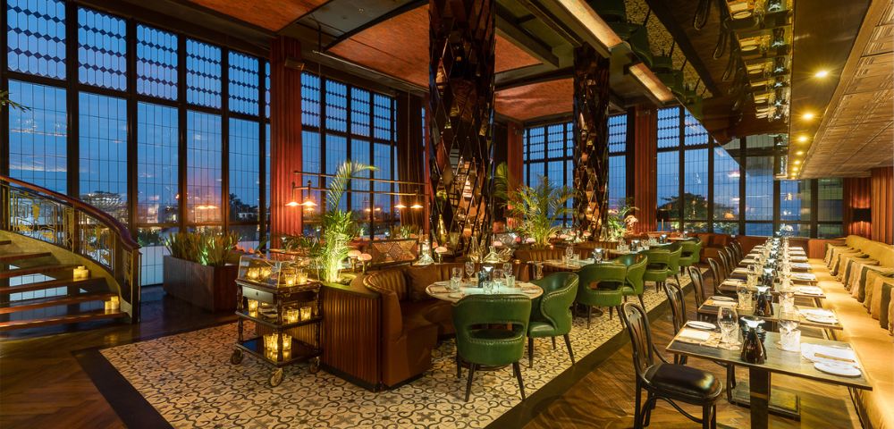 Luxurious restaurant interior with elegant green chairs, wooden tables, and warm lighting. Floor-to-ceiling windows provide city night views, enhancing the cozy ambiance.