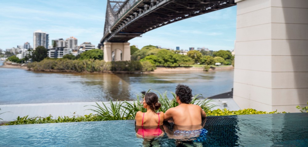 Two people relax in an infinity pool, facing a large steel bridge spanning across a river. The backdrop includes city buildings and lush greenery.