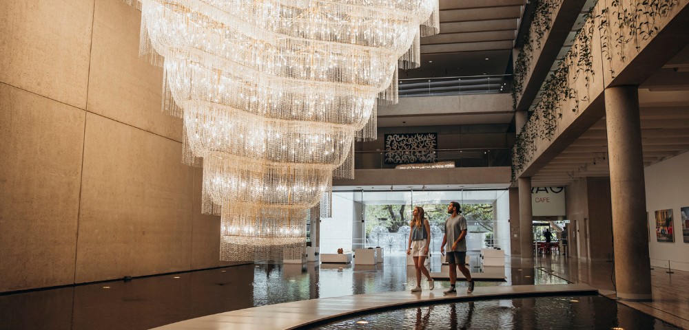 A grand chandelier with cascading lights illuminates an elegant, modern lobby with high ceilings. Two people walk along a path above a reflecting pool.