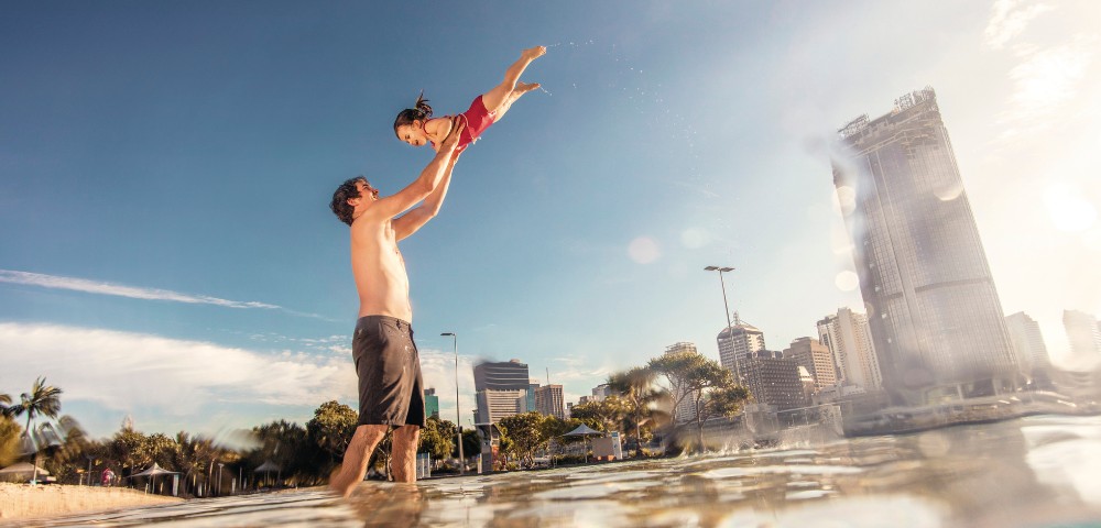A man in swim trunks joyfully lifts a laughing child in a swimsuit above shallow water. The cityscape, palm trees, and clear sky create a vibrant, carefree scene.