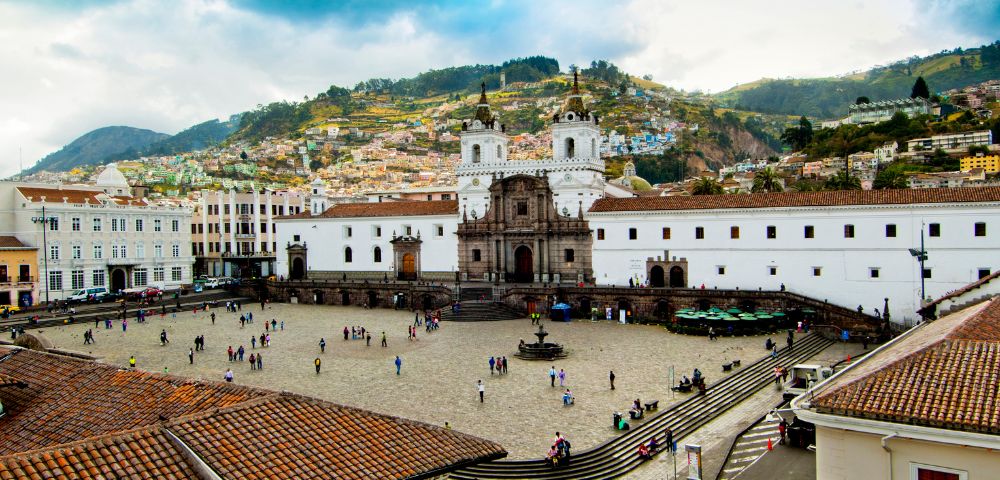 A bustling plaza in front of an ornate colonial church with twin bell towers in Quito, surrounded by colorful hillside houses, under a cloudy sky.