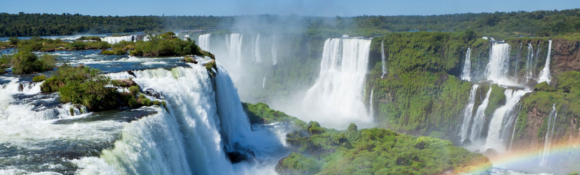 Spectacular view of Iguazu Falls with cascading waters, lush greenery, mist, and a vibrant rainbow. The scene conveys awe and natural beauty.