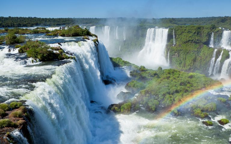 Spectacular view of Iguazu Falls with cascading waters, lush greenery, mist, and a vibrant rainbow. The scene conveys awe and natural beauty.
