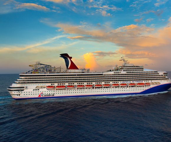 A large cruise ship sails on a calm ocean under a vibrant sunset sky. The ship features multiple decks and a distinctive red funnel.