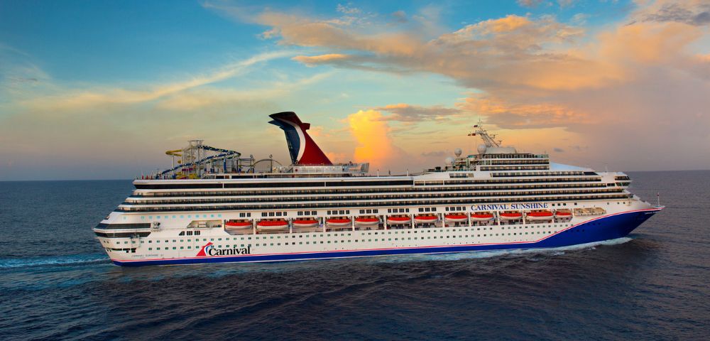 A large cruise ship sails on a calm ocean under a vibrant sunset sky. The ship features multiple decks and a distinctive red funnel.