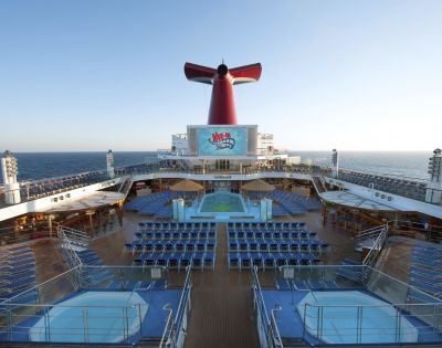 The image shows the deck of a cruise ship with rows of blue lounge chairs facing a large pool and red funnel. The sea and clear sky are in the background.