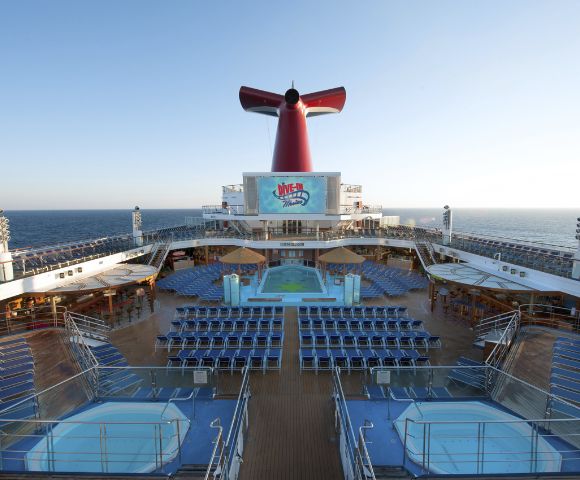 The image shows the deck of a cruise ship with rows of blue lounge chairs facing a large pool and red funnel. The sea and clear sky are in the background.