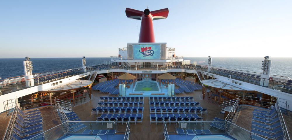 The image shows the deck of a cruise ship with rows of blue lounge chairs facing a large pool and red funnel. The sea and clear sky are in the background.