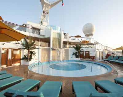 Sunny cruise ship deck with a round pool and cascading waterfall. Surrounded by palm plants and turquoise loungers, there's a relaxed, luxurious vibe.