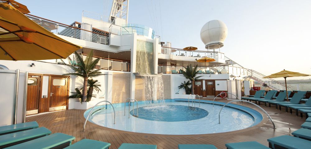 Sunny cruise ship deck with a round pool and cascading waterfall. Surrounded by palm plants and turquoise loungers, there's a relaxed, luxurious vibe.