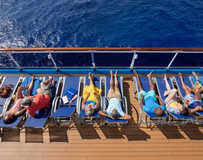 Six people sunbathe on lounge chairs aboard a cruise ship, overlooking a deep blue ocean. The scene conveys relaxation and leisure.