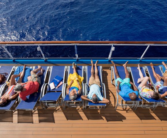 Six people sunbathe on lounge chairs aboard a cruise ship, overlooking a deep blue ocean. The scene conveys relaxation and leisure.
