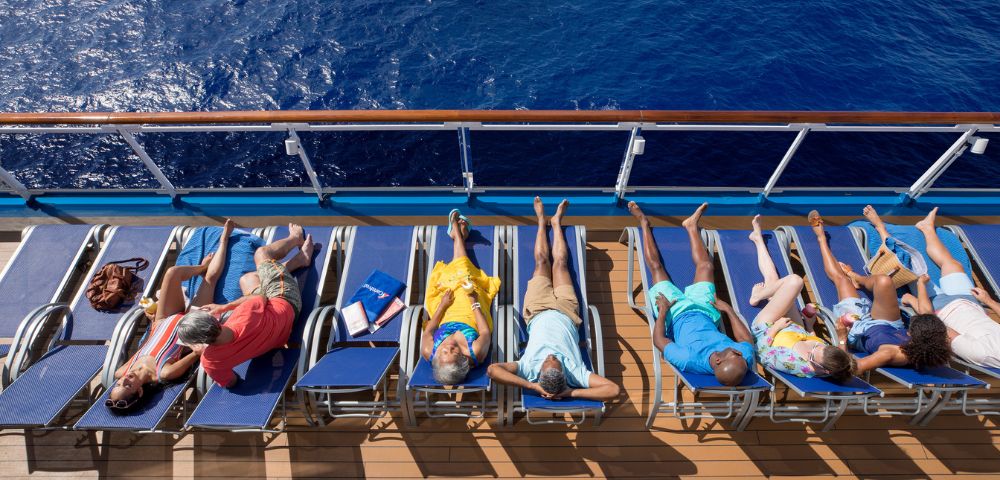 Six people sunbathe on lounge chairs aboard a cruise ship, overlooking a deep blue ocean. The scene conveys relaxation and leisure.