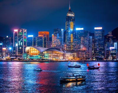 Nighttime cityscape of Hong Kong's vibrant skyline, illuminated skyscrapers reflecting on shimmering water, with traditional boats adding cultural charm.