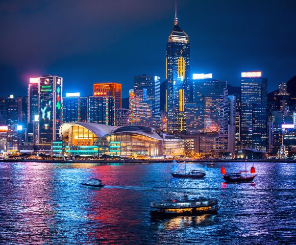 Nighttime cityscape of Hong Kong's vibrant skyline, illuminated skyscrapers reflecting on shimmering water, with traditional boats adding cultural charm.