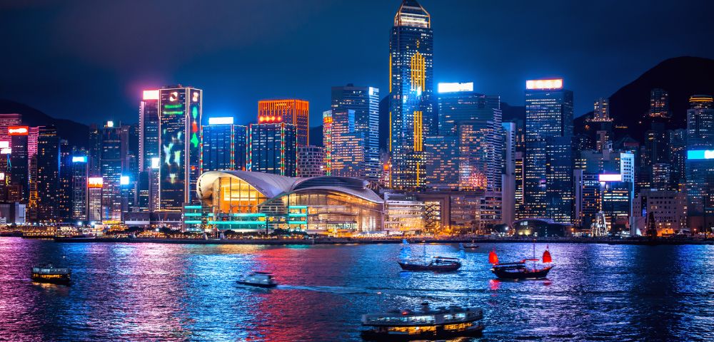 Nighttime cityscape of Hong Kong's vibrant skyline, illuminated skyscrapers reflecting on shimmering water, with traditional boats adding cultural charm.