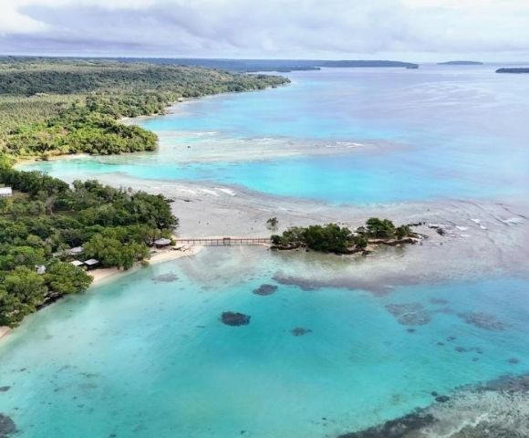 Aerial view of a serene coastline with turquoise waters, lush green forest, a small bridge connecting islands, and a cloudy sky above. Peaceful and tropical.