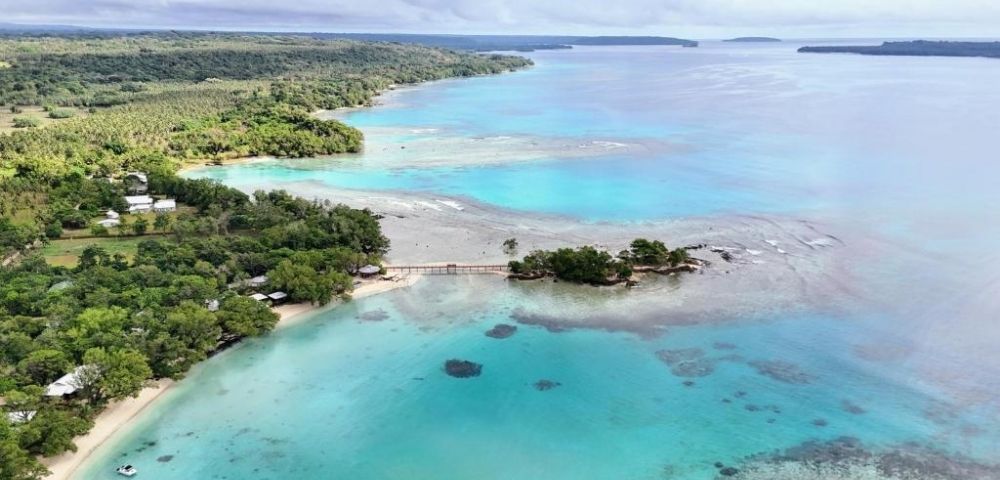 Aerial view of a serene coastline with turquoise waters, lush green forest, a small bridge connecting islands, and a cloudy sky above. Peaceful and tropical.