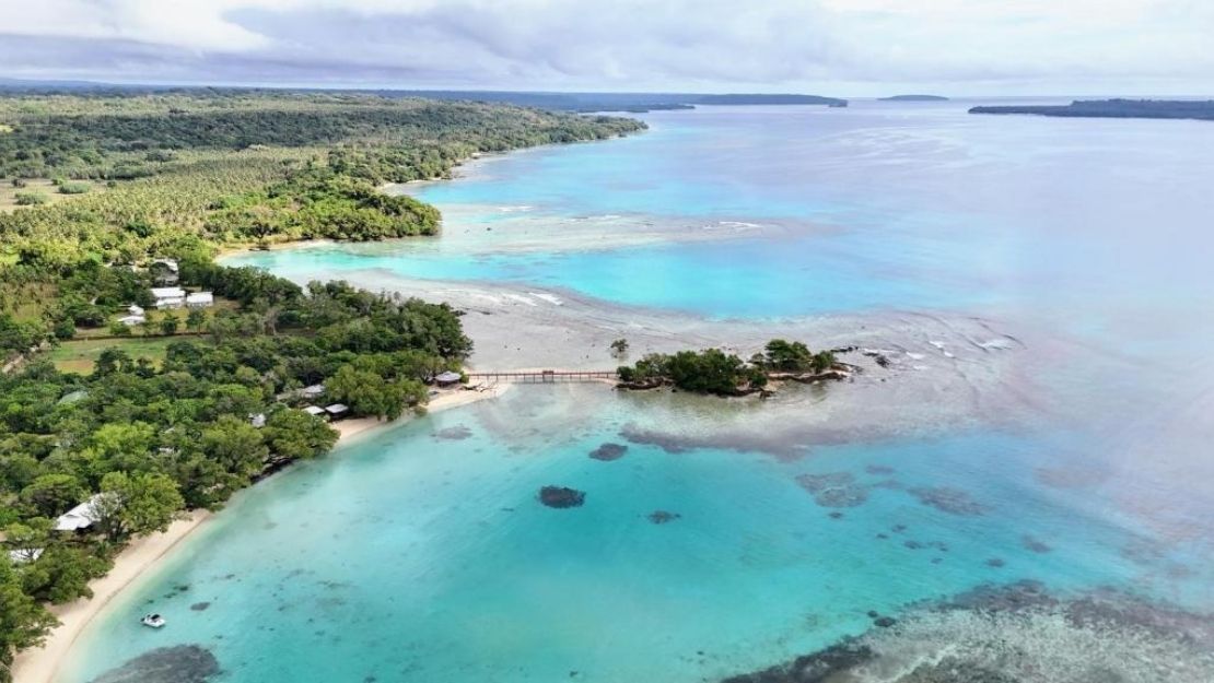 Aerial view of a serene coastline with turquoise waters, lush green forest, a small bridge connecting islands, and a cloudy sky above. Peaceful and tropical.