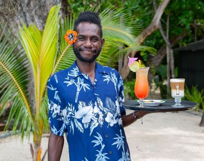 Man in a blue tropical shirt, smiling, holds a tray with colorful cocktails. He's standing on a sandy beach with palm trees in the background.