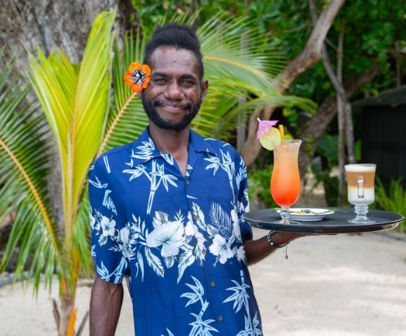 Man in a blue tropical shirt, smiling, holds a tray with colorful cocktails. He's standing on a sandy beach with palm trees in the background.