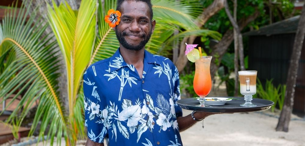 Man in a blue tropical shirt, smiling, holds a tray with colorful cocktails. He's standing on a sandy beach with palm trees in the background.