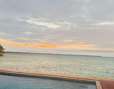 Tranquil ocean scene at sunset with cloudy sky, gentle waves, and pastel colors. A pool in the foreground enhances the serene atmosphere.