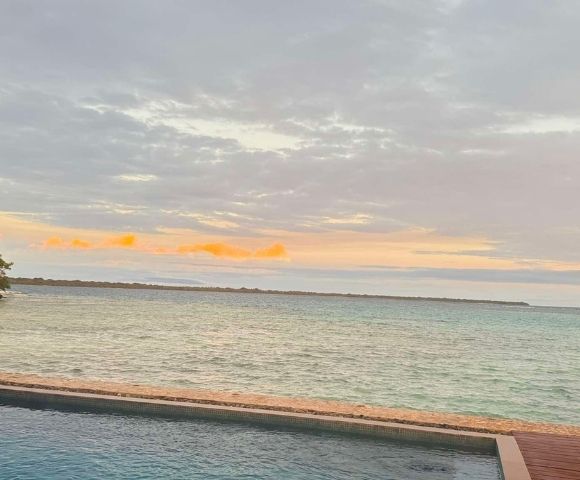 Tranquil ocean scene at sunset with cloudy sky, gentle waves, and pastel colors. A pool in the foreground enhances the serene atmosphere.
