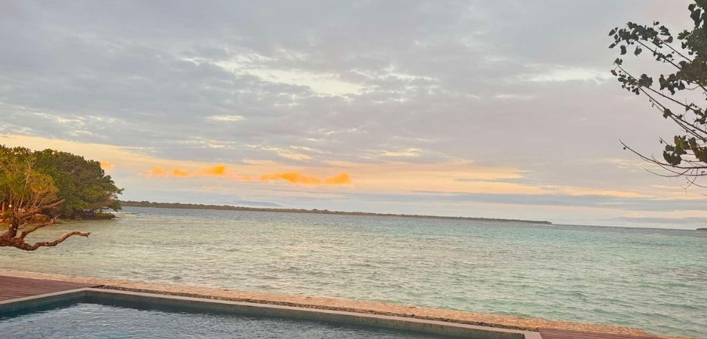Tranquil ocean scene at sunset with cloudy sky, gentle waves, and pastel colors. A pool in the foreground enhances the serene atmosphere.