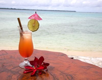 A tropical drink with a pink umbrella and lime slice sits on a wooden table beside the beach. Red flowers add a vibrant touch, evoking a relaxed, vacation vibe.