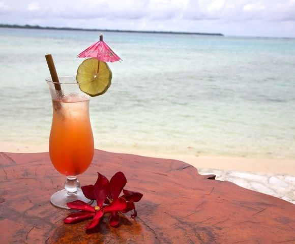 A tropical drink with a pink umbrella and lime slice sits on a wooden table beside the beach. Red flowers add a vibrant touch, evoking a relaxed, vacation vibe.