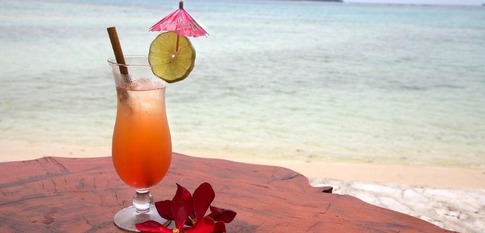 A tropical drink with a pink umbrella and lime slice sits on a wooden table beside the beach. Red flowers add a vibrant touch, evoking a relaxed, vacation vibe.