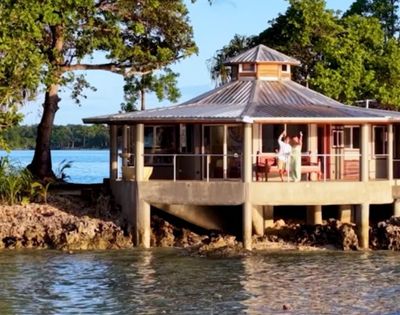 A round, elevated beach house with large windows and a metal roof sits by the water. A person waves from the deck, surrounded by lush trees and a calm sea.