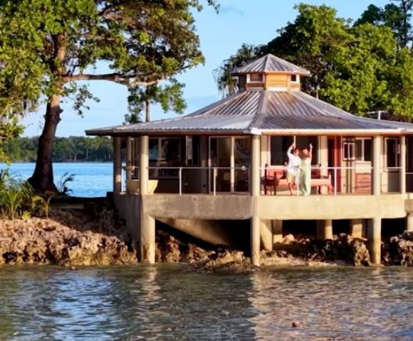 A round, elevated beach house with large windows and a metal roof sits by the water. A person waves from the deck, surrounded by lush trees and a calm sea.