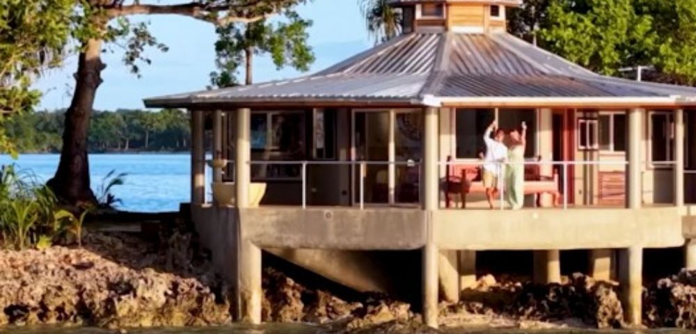 A round, elevated beach house with large windows and a metal roof sits by the water. A person waves from the deck, surrounded by lush trees and a calm sea.