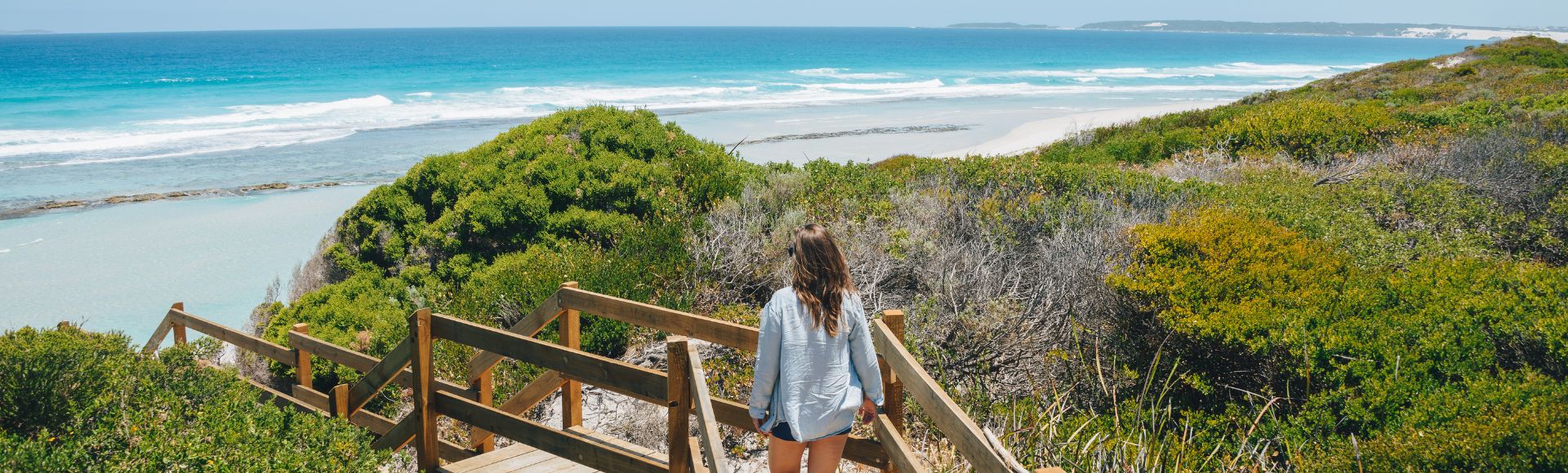 A woman walks down a wooden path toward a pristine beach with turquoise waves. Lush green bushes line the path, conveying a serene, inviting atmosphere.