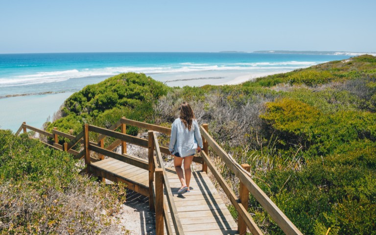 A woman walks down a wooden path toward a pristine beach with turquoise waves. Lush green bushes line the path, conveying a serene, inviting atmosphere.
