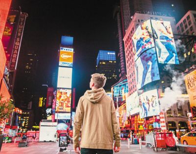 A person in a tan hoodie stands in Times Square at night, surrounded by bright neon billboards and city lights, conveying a sense of awe and wonder.