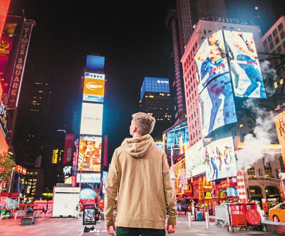 A person in a tan hoodie stands in Times Square at night, surrounded by bright neon billboards and city lights, conveying a sense of awe and wonder.