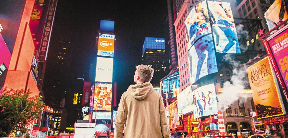 A person in a tan hoodie stands in Times Square at night, surrounded by bright neon billboards and city lights, conveying a sense of awe and wonder.