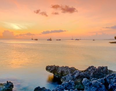 Tranquil sunset over a calm ocean, with vibrant orange and pink sky reflections on the water. Rocky foreground and distant sailboats create a serene scene.
