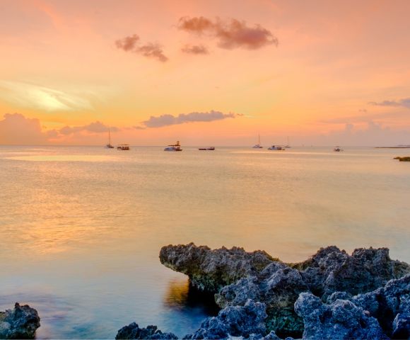 Tranquil sunset over a calm ocean, with vibrant orange and pink sky reflections on the water. Rocky foreground and distant sailboats create a serene scene.