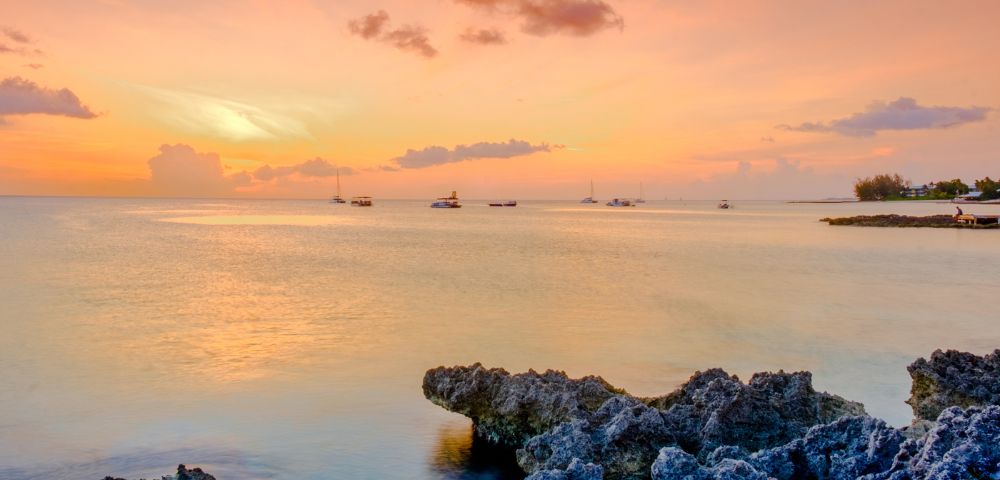 Tranquil sunset over a calm ocean, with vibrant orange and pink sky reflections on the water. Rocky foreground and distant sailboats create a serene scene.