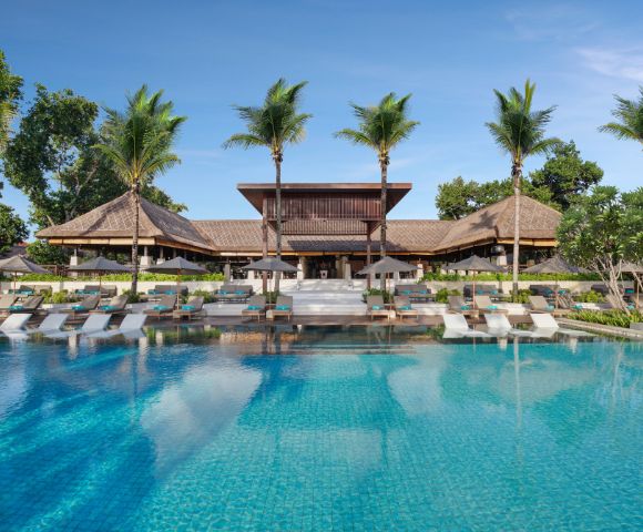 Luxurious tropical resort with a large, clear blue pool, surrounded by palm trees and wooden architecture, creating a serene and inviting atmosphere.