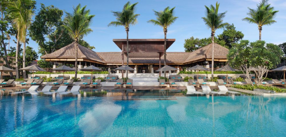 Luxurious tropical resort with a large, clear blue pool, surrounded by palm trees and wooden architecture, creating a serene and inviting atmosphere.
