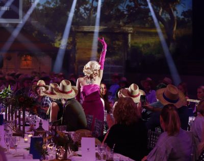 A performer in a vibrant pink dress with matching glove sings amid a lively audience seated at candlelit tables, beneath dramatic stage lighting.